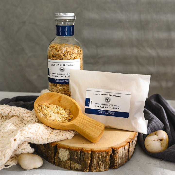 Wooden scoop filled with Edie Herbal Bath Soak next to full pouch and glass bottle on wooden slab next to a cream colored knit washcloth, grey towel and pebbles.