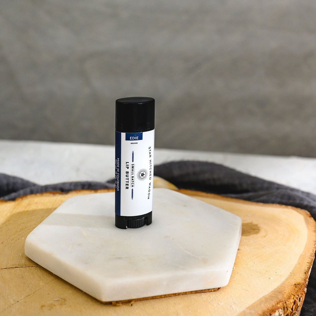 A black tube of lip butter, placed on a stone and wooden surface with a grey cloth in the background.