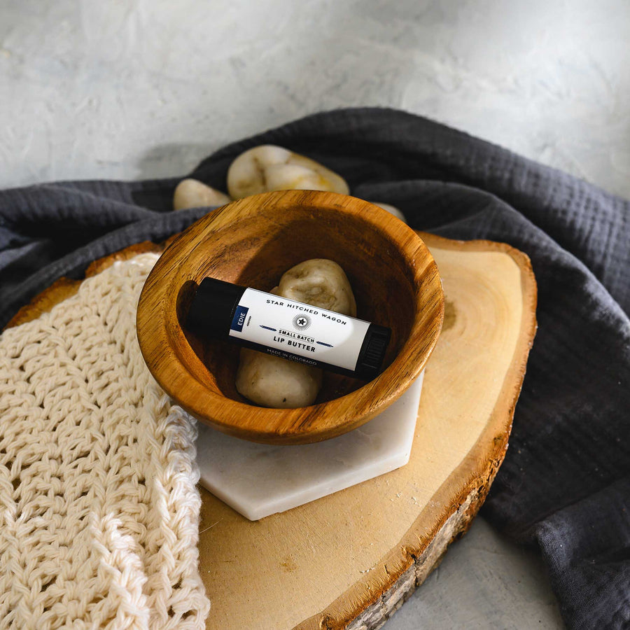 A small wooden bowl containing a black tube of lip butter, placed on a wooden surface with a grey cloth and some stones in the background.