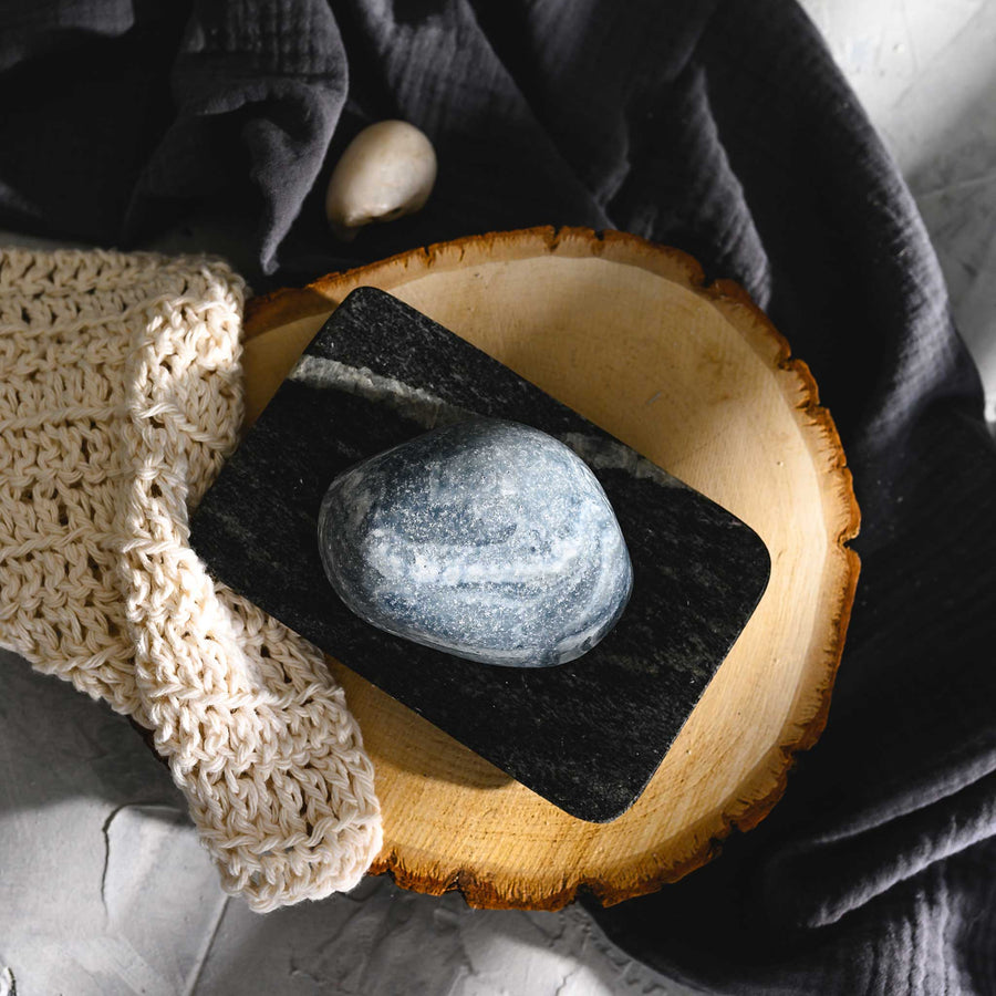 Edie river stone salt soap bar resting on a wooden surface with a grayish-blue pattern, accompanied by a beige textured cloth.