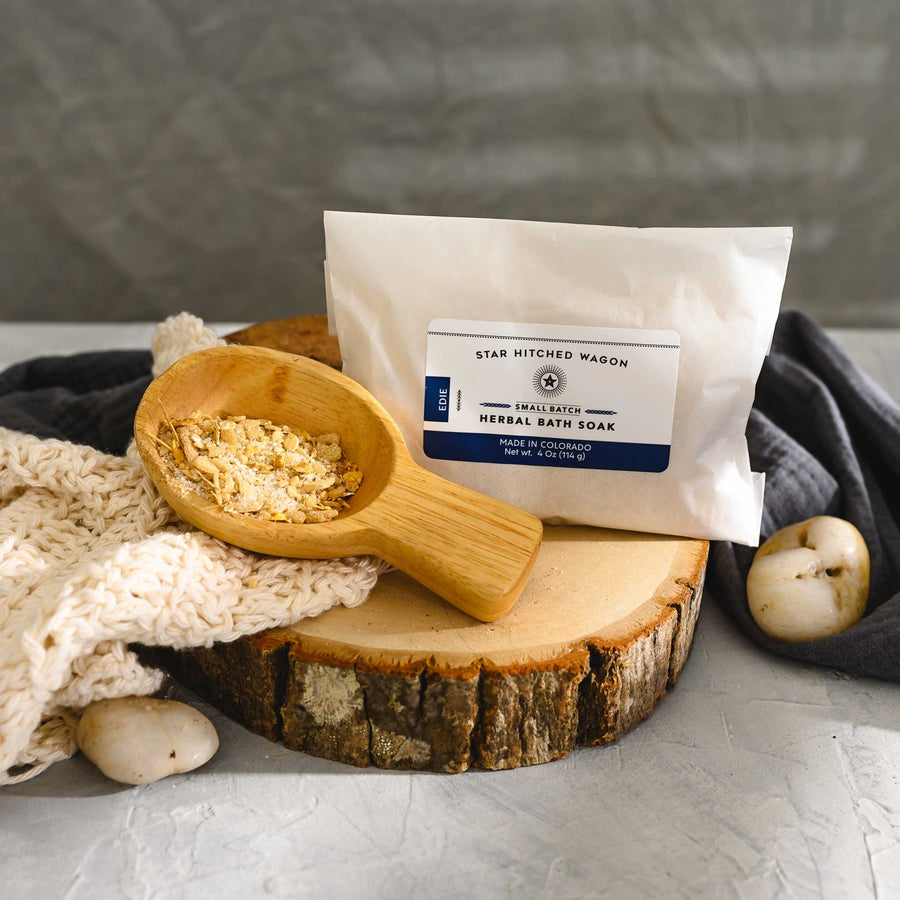 Wooden scoop filled with Edie Herbal Bath Soak next to full pouch on wooden slab next to a cream colored knit washcloth.