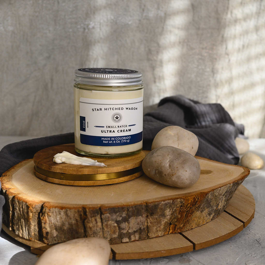 Edie Ultra Cream in a glass jar with an aluminum screw top lid, displayed on a wooden surface with a grey cloth and pebbles in the background.
