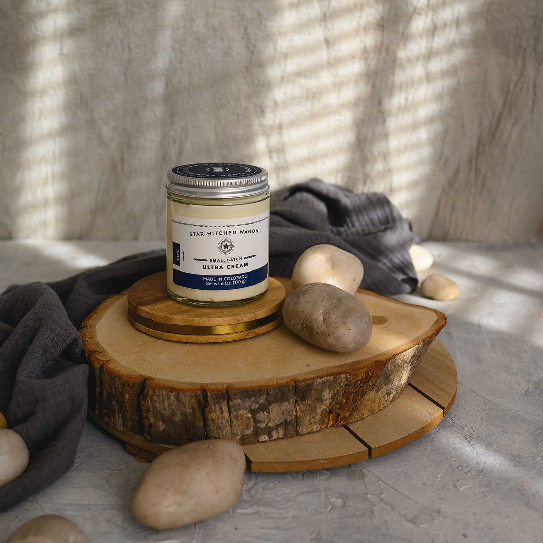 Edie Ultra Cream in a glass jar with an aluminum screw top lid, displayed on a wooden surface with a grey cloth and pebbles in the background.