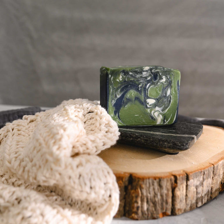 Edie essential oil soap bar with swirls of navy blue, green, and white, placed on a wooden surface with a knit washcloth beside it.