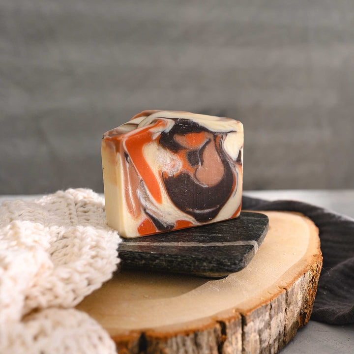 Nora essential oil soap bar with a swirl pattern in shades of orange, brown, and white, placed on a stone and wooden surface next to a crocheted washcloth with a grey towel in the background.