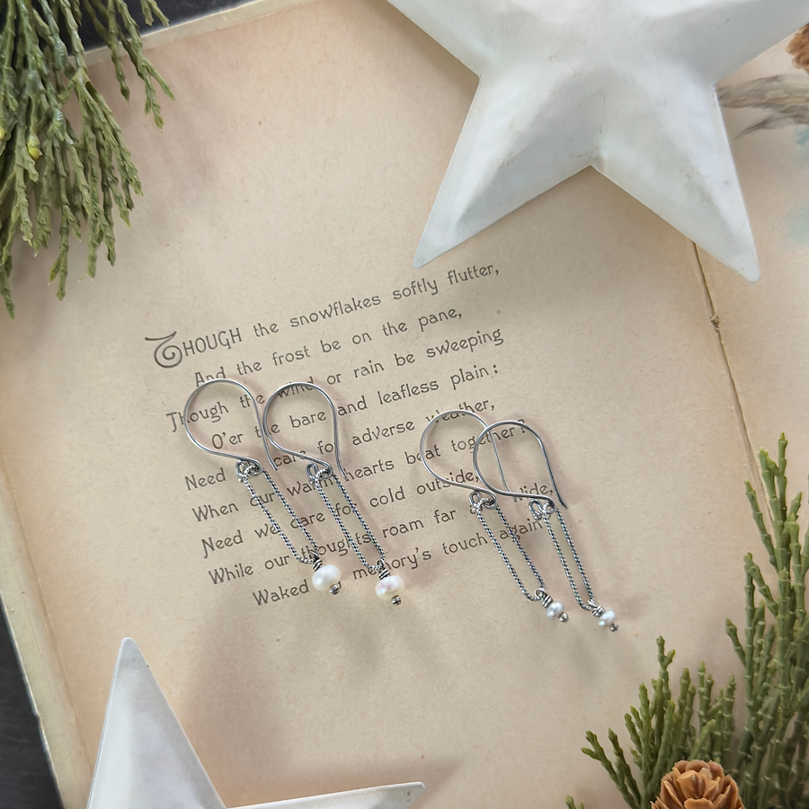 Two pairs of oxidized sterling silver pearl drop earrings displayed on an open vintage book with winter greenery and white star decor.