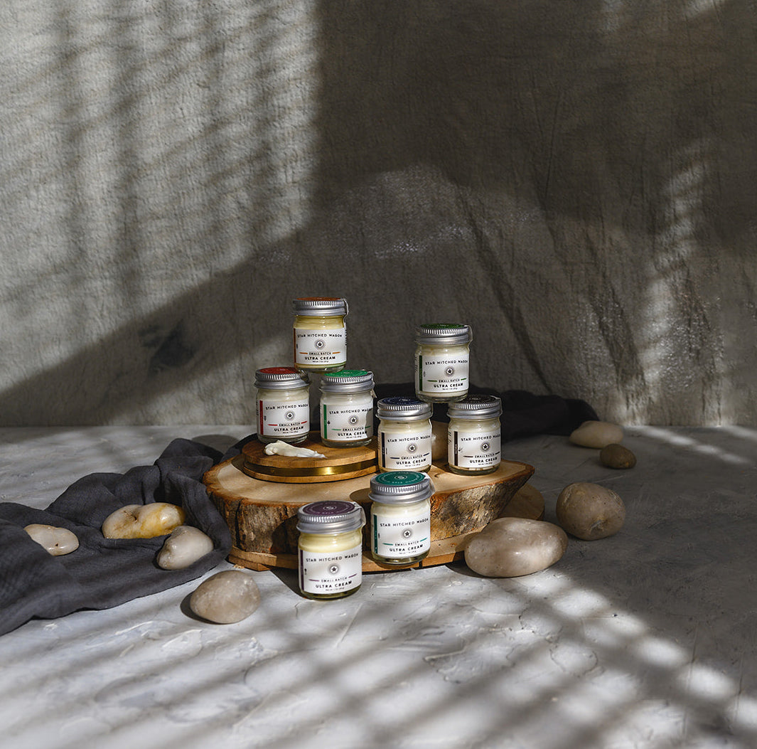 A styled product shot of full collection of 1 ounce ultra cream jars arranged on a wooden slice with stones and a dark cloth against a textured wall, lit by dramatic window shadows.