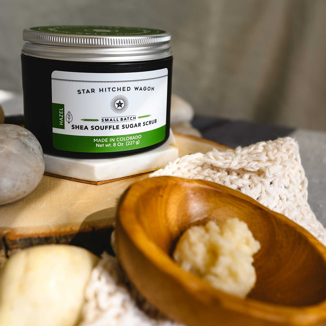 An 8 ounce jar of Hazel Shea Souffle Sugar Scrub is displayed on a wooden surface with a wooden spoon resting on a knit cloth, surrounded by small river stones.