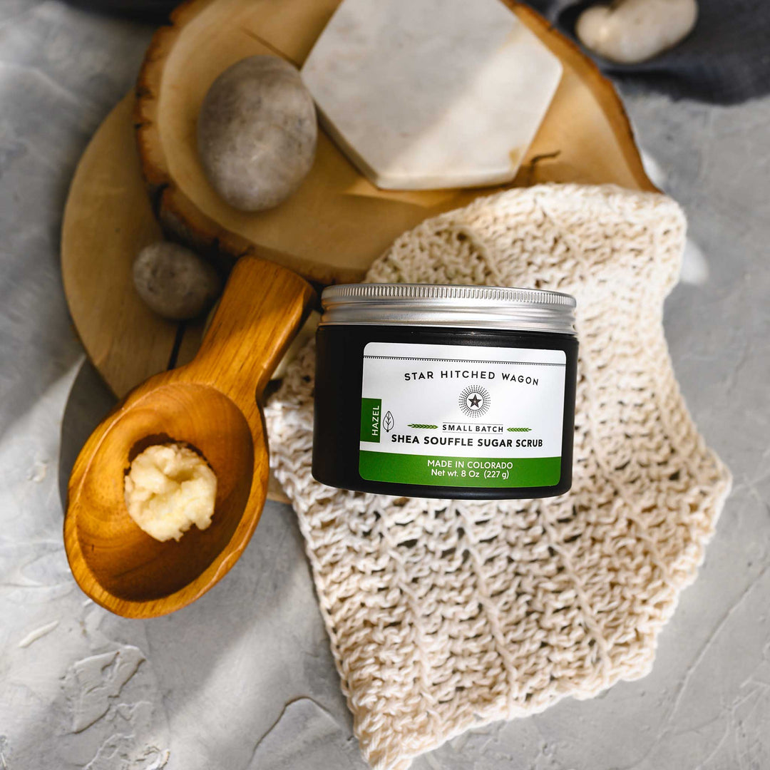 An 8 ounce jar of Hazel Shea Souffle Sugar Scrub is displayed on a wooden surface with a wooden spoon resting on a knit cloth, surrounded by small river stones.