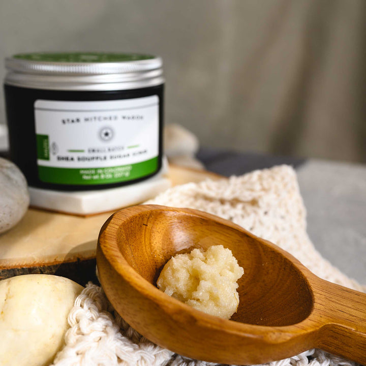 A close up of Hazel Shea Soufflé Sugar Scrub in a wooden scoop displayed on a wooden surface with a knit cloth with a full jar in the background along with small river stones.