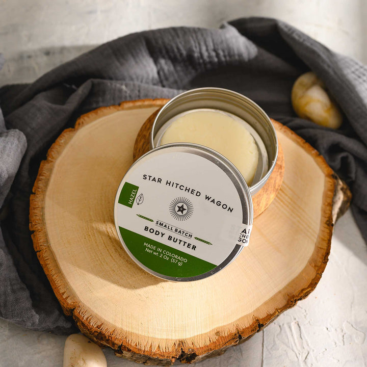 Hazel body butter moisturizer in reusable tin, handcrafted with organic oils and butters to nourish and protect dry skin, set on a wooden slab with pebbles and grey towel in background.