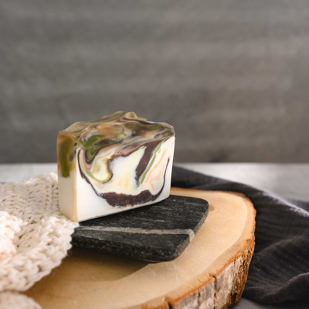 Hazel essential oil soap bar with swirls of peach, green, brown, and cream, placed on a wooden surface with a knit washcloth beside it.