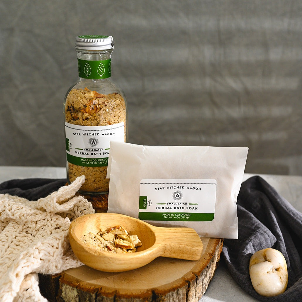 Wooden scoop filled with Hazel Herbal Bath Soak next to full pouch and glass bottle on wooden slab next to a cream colored knit washcloth, a grey towel and pebbles.
