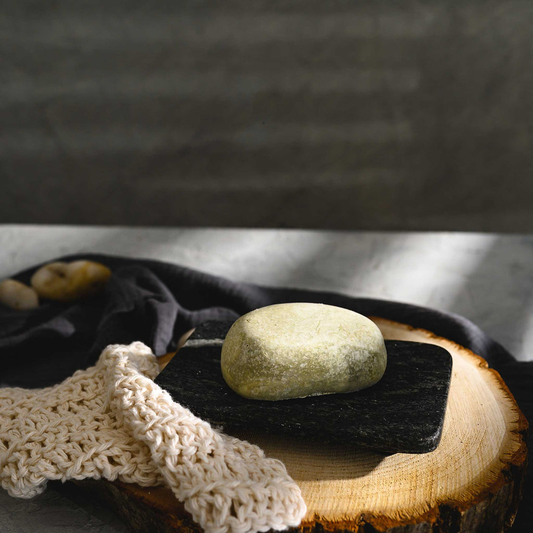 Hazel river stone salt soap bar, placed on a wooden surface with a dark cloth and a knit beige soap dish in the foreground.
