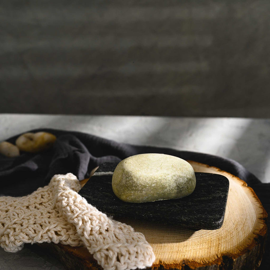 Hazel river stone salt soap bar, placed on a wooden surface with a dark cloth and a knit beige soap dish in the foreground.