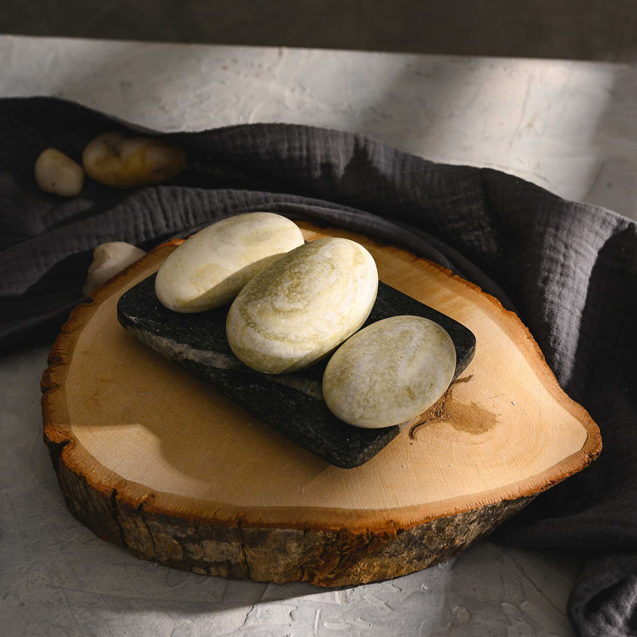 Hazel river stone salt soap trio with a smooth, matte finish, displayed on a wooden slice, with a dark cloth in the background.