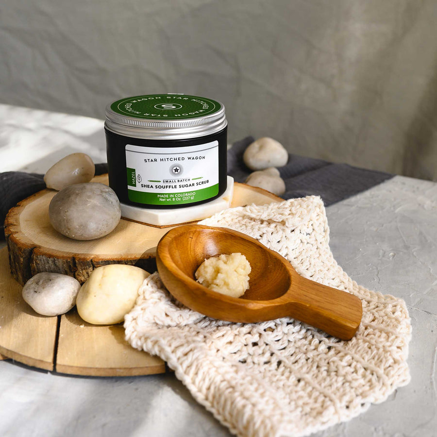 An 8 ounce jar of Hazel Shea Souffle Sugar Scrub is displayed on a wooden surface with a wooden spoon resting on a knit cloth, surrounded by small river stones.