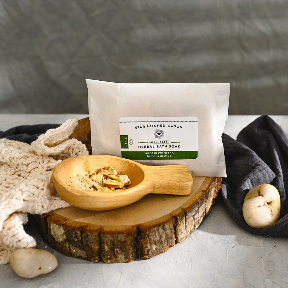 Wooden scoop filled with Hazel Herbal Bath Soak next to full pouch on wooden slab next to a cream colored knit washcloth, a grey towel and pebbles.