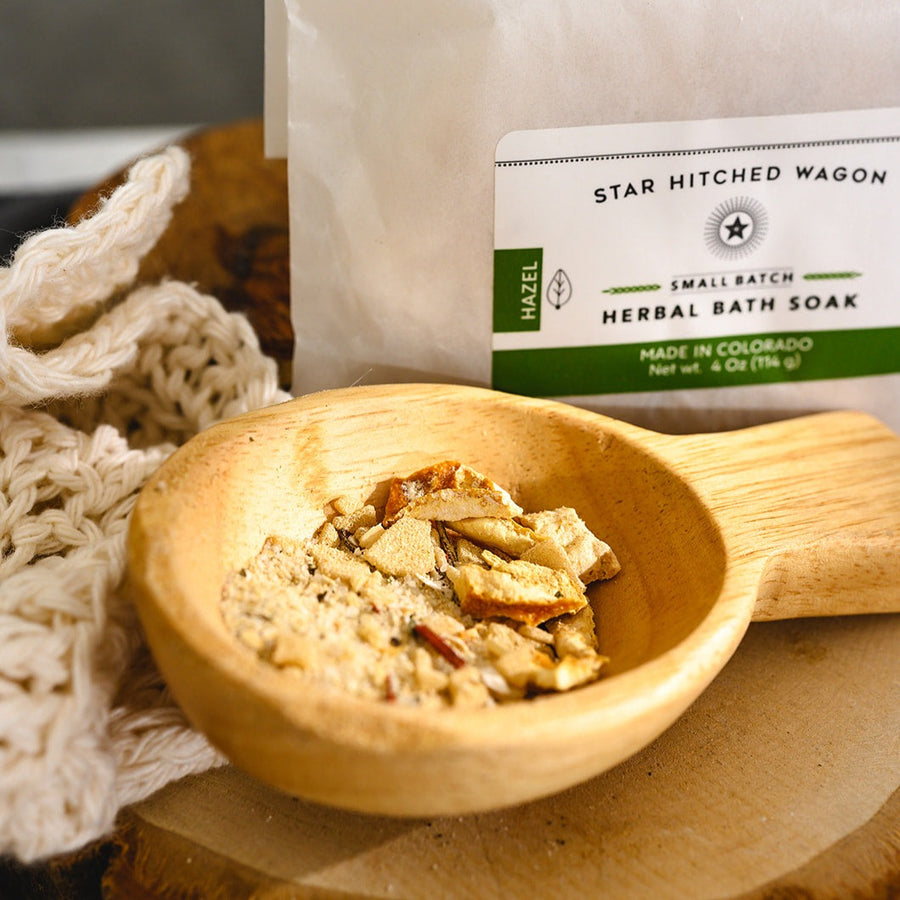 Wooden scoop filled with Hazel Herbal Bath Soak next to full pouch on wooden slab next to a cream colored knit washcloth.