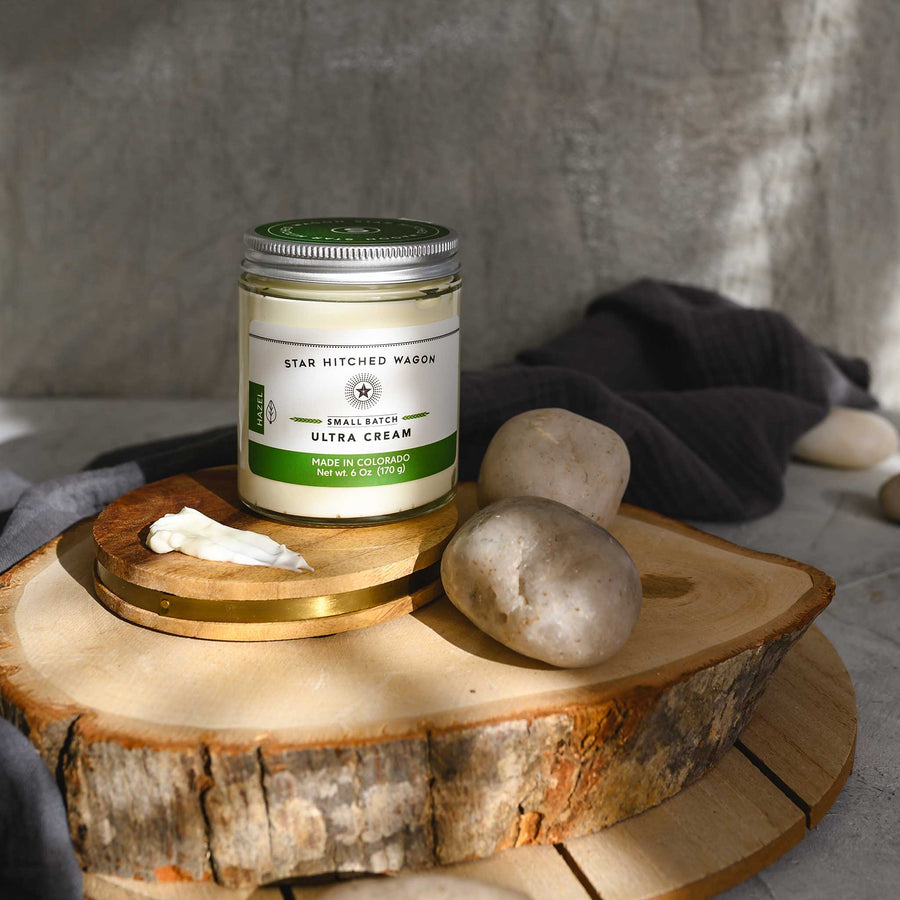Hazel Ultra Cream in a glass jar with an aluminum screw top lid, placed on a wooden surface with pebbles and grey towel in the background.