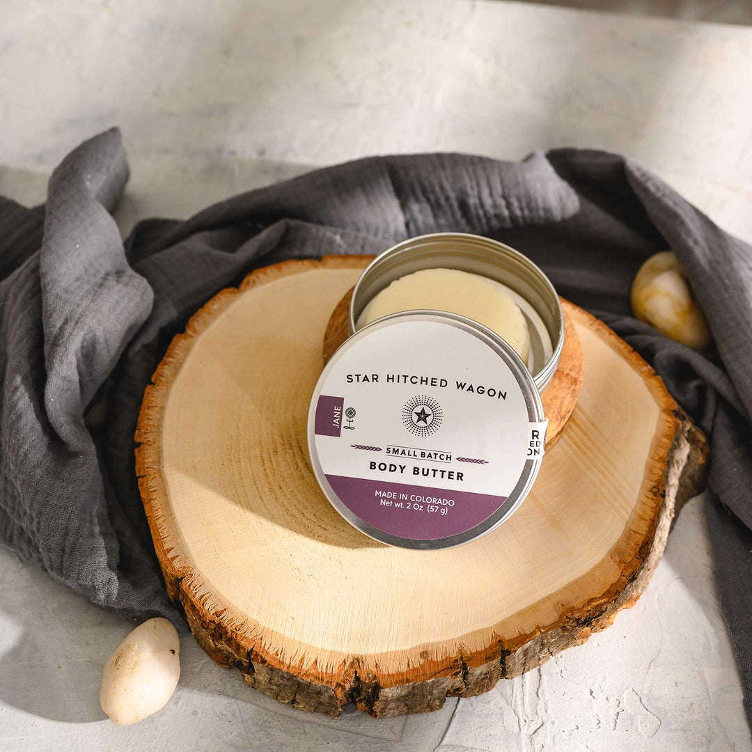 Jane 2 Ounce Body Butter Moisturizer Bar in reusable tin on wood and stone surface with pebbles and grey towel in background