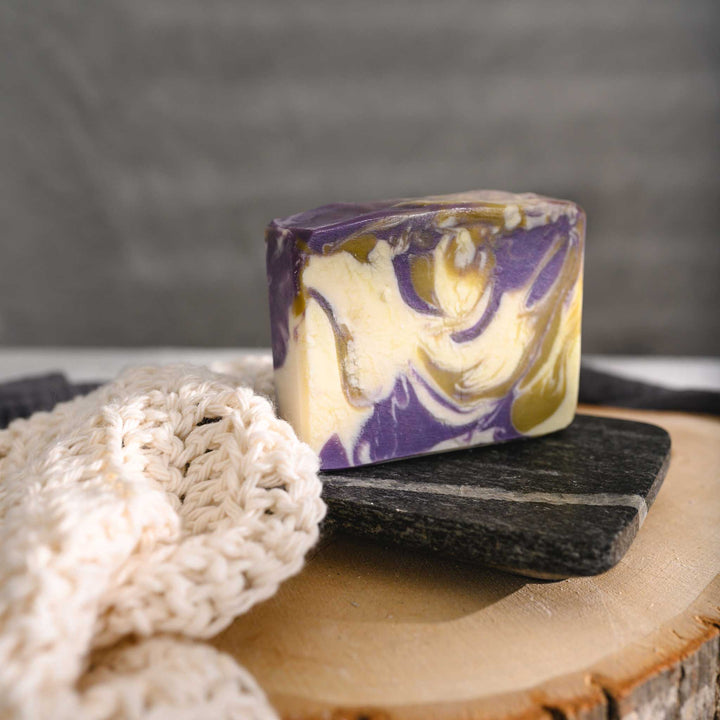 Jane essential oil soap bar with swirls of purple, green, and cream, resting on a stone and wooden surface next to a cream colored knit washcloth with a grey towel in the background.