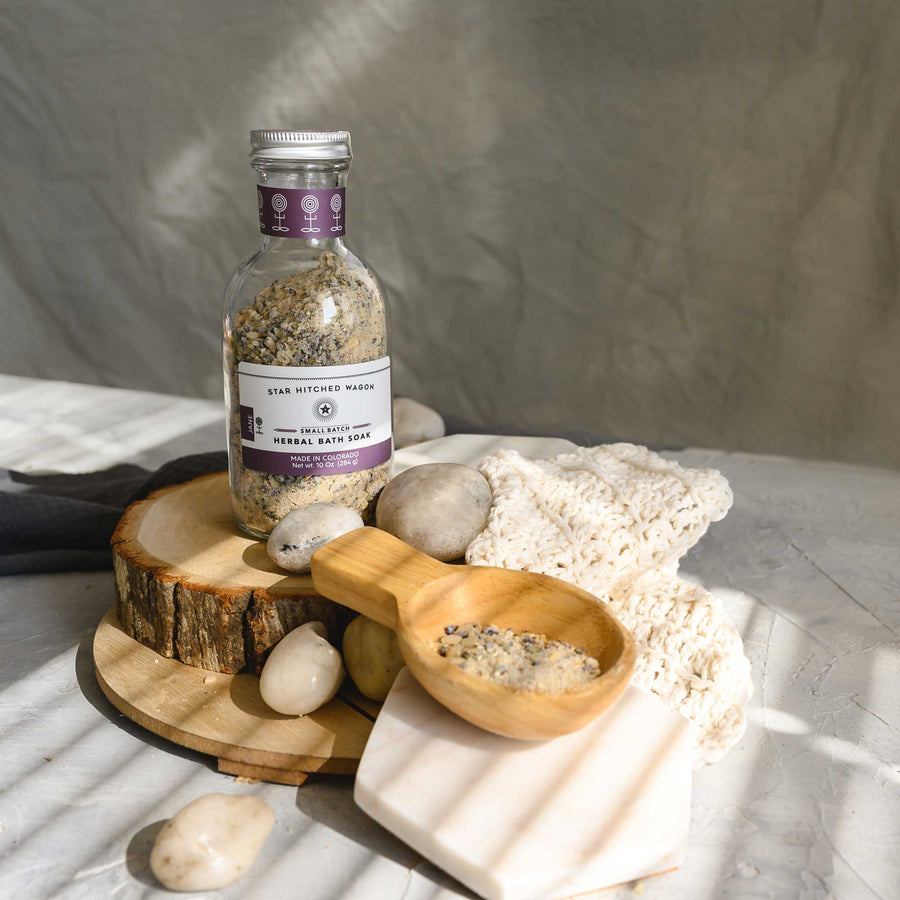 Jane Herbal Bath Soak in a glass bottle next to a wooden spoon filled with the soak and a cream colored wash cloth and river stones.