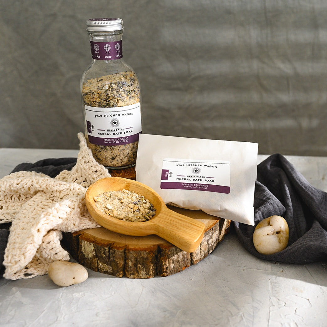 Wooden scoop filled with Jane Herbal Bath Soak next to full pouch and glass bottle on wooden slab next to a cream colored knit washcloth, a grey towel and pebbles.