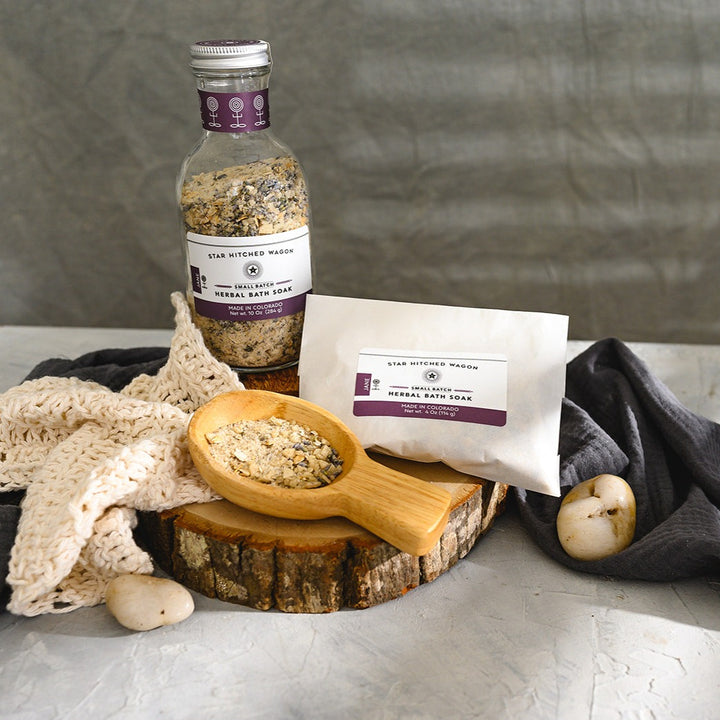 Wooden scoop filled with Jane Herbal Bath Soak next to full pouch and glass bottle on wooden slab next to a cream colored knit washcloth, a grey towel and pebbles.