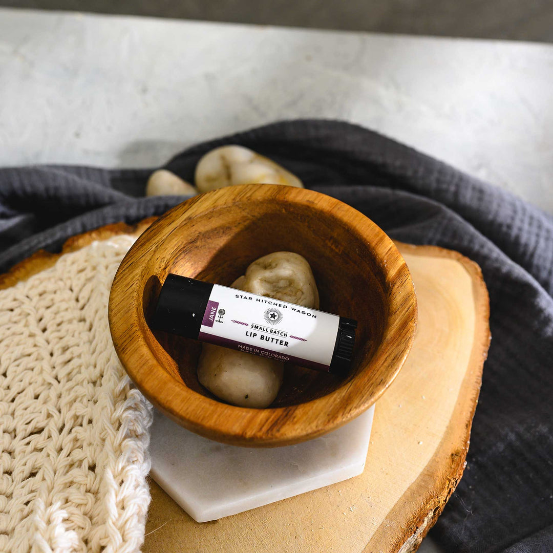A small Wooden bowl with a tube of Jane lip butter, placed on a wooden surface, alongside a knit washcloth, a grey towel and pebbles. 