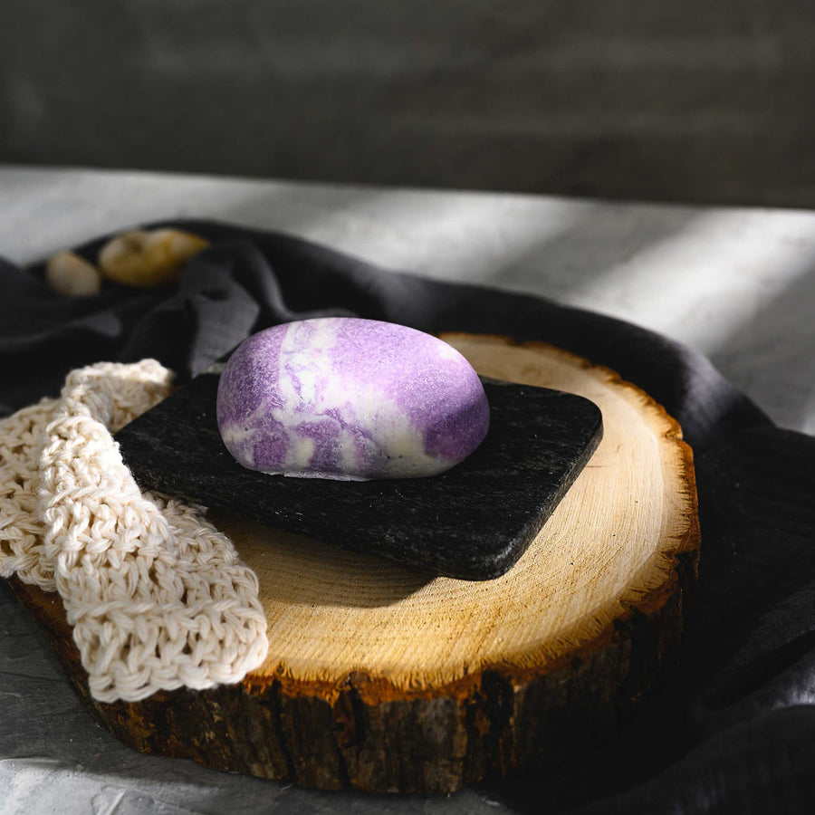 Jane River Stone Salt Soap Bar with a marbled pattern of purple and white, placed on a wooden slice next to a crocheted wash cloth on a black soap rest, with a dark cloth in the background.