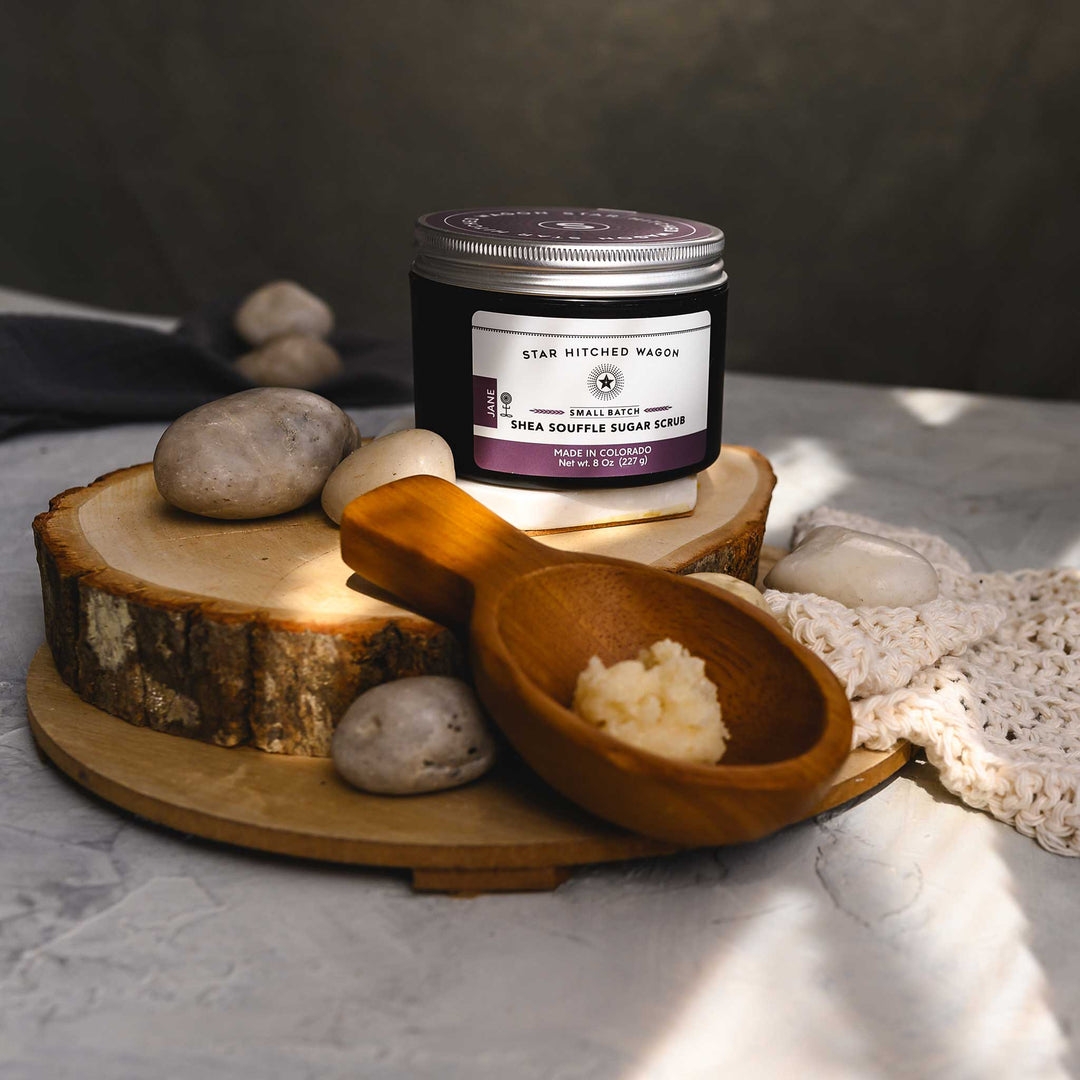 A jar of Jane 8 Ounce Shea Soufflé Sugar Scrub is displayed on a wooden slice, alongside a wooden scoop and a knit washcloth, with a few stones around. The setup is on a marble surface.