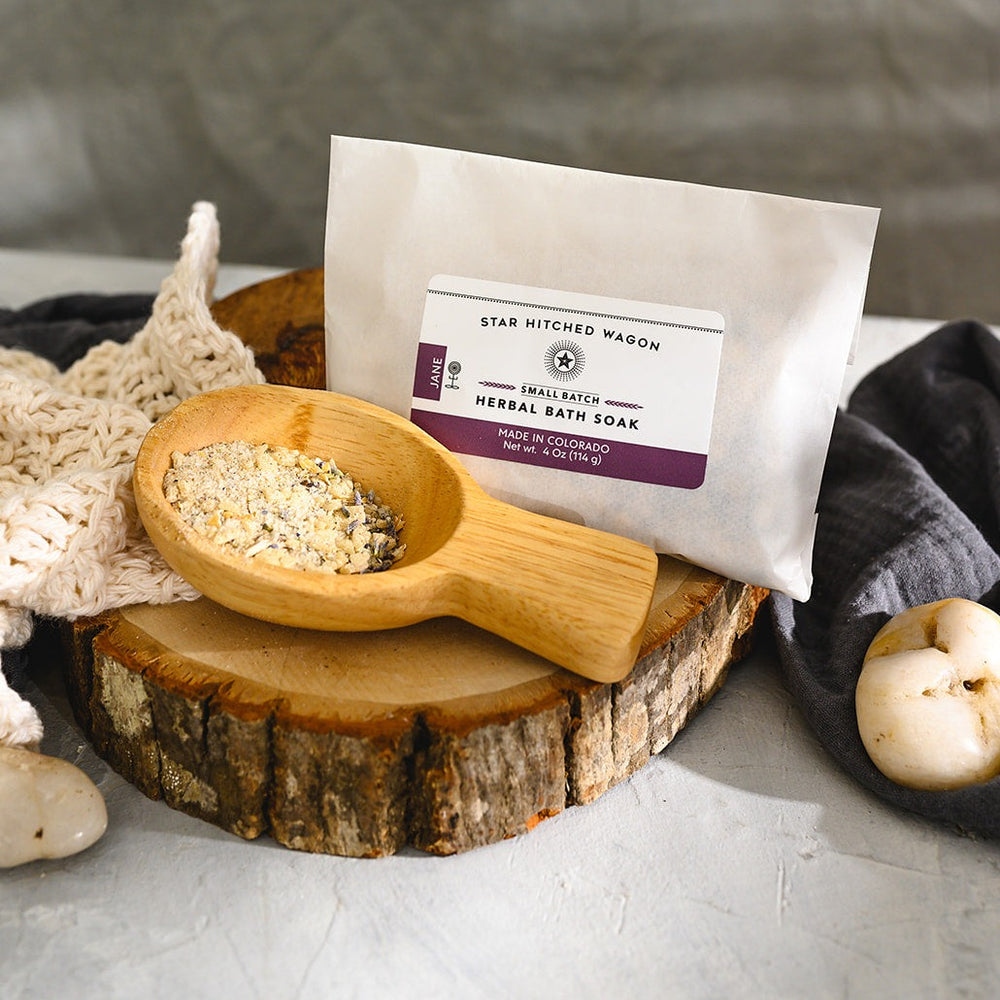 Wooden scoop filled with Jane Herbal Bath Soak next to full pouch on wooden slab next to a cream colored washcloth, a grey towel and pebbles.