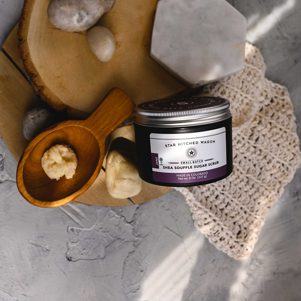 A jar of Jane 8 Ounce Shea Soufflé Sugar Scrub is displayed on a wooden slice, alongside a wooden scoop and a knit washcloth, with a few stones around. The setup is on a marble surface.