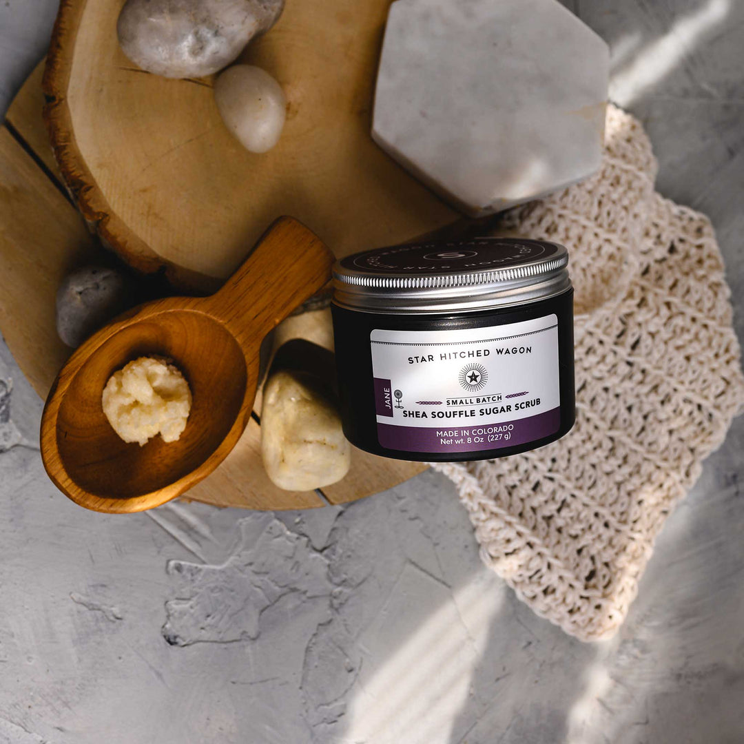 A jar of Jane 8 Ounce Shea Soufflé Sugar Scrub is displayed on a wooden slice, alongside a wooden scoop and a knit washcloth, with a few stones around. The setup is on a marble surface.