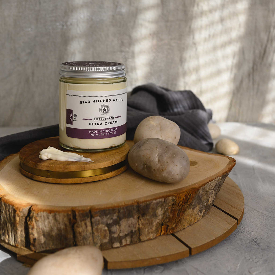 Jane Ultra Cream in a glass jar placed on a wooden disc, alongside some stones and a grey towel. 