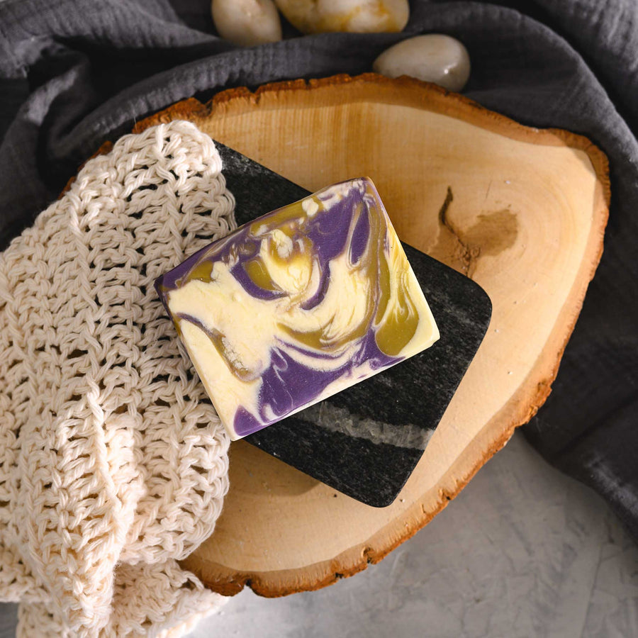 Jane essential oil soap bar with swirls of purple, yellow, and beige, resting on a wooden surface with a cream colored knit washcloth in the background.