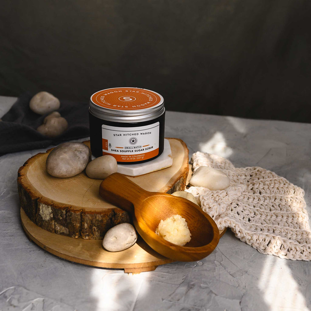 A jar of Jess 8 Ounce Shea Soufflé Sugar Scrub is displayed on a wooden surface, alongside a wooden spoon, a knit washcloth and small river stones.