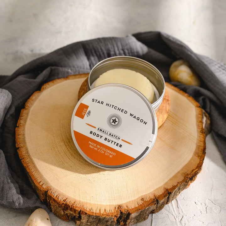 Jess body butter moisturizer in reusable tin, made with organic oils and butters to nourish dry skin. Scented with tangerine and patchouli. Set on a wooden and stone surface with pebbles and grey towel in the background.