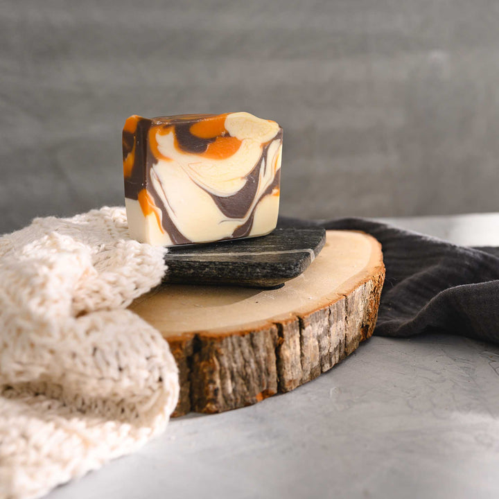 Jess essential oil soap bar with a swirl pattern of orange, brown, and cream colors, placed on a stone and wood surface with a cream colored knitted washcloth beside it and a grey towel in the background.