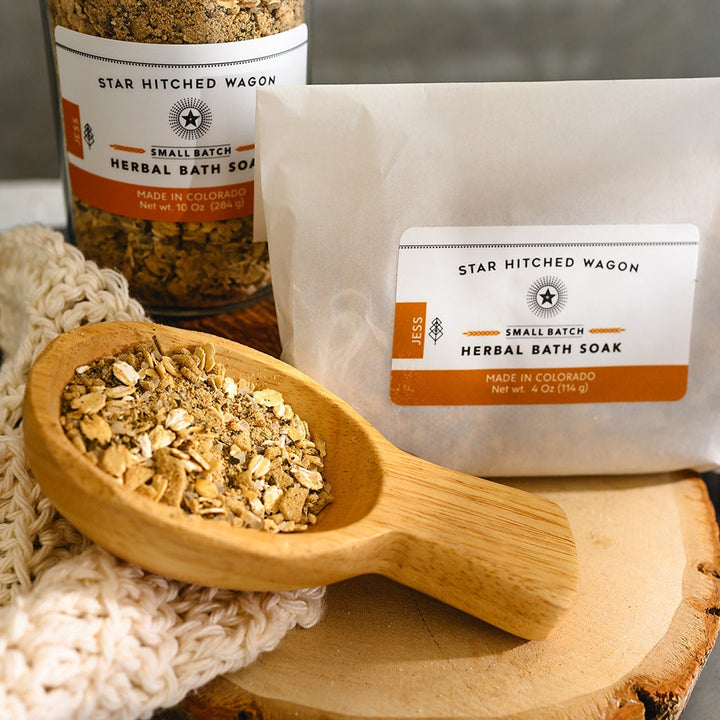 Wooden scoop filled with Kate Herbal Bath Soak next to full pouch and glass bottle on wooden slab next to a cream colored knit washcloth.