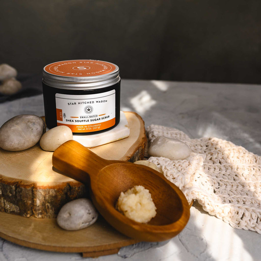 A jar of Jess 8 Ounce Shea Soufflé Sugar Scrub is displayed on a wooden surface, alongside a wooden spoon, a knit washcloth and small river stones.