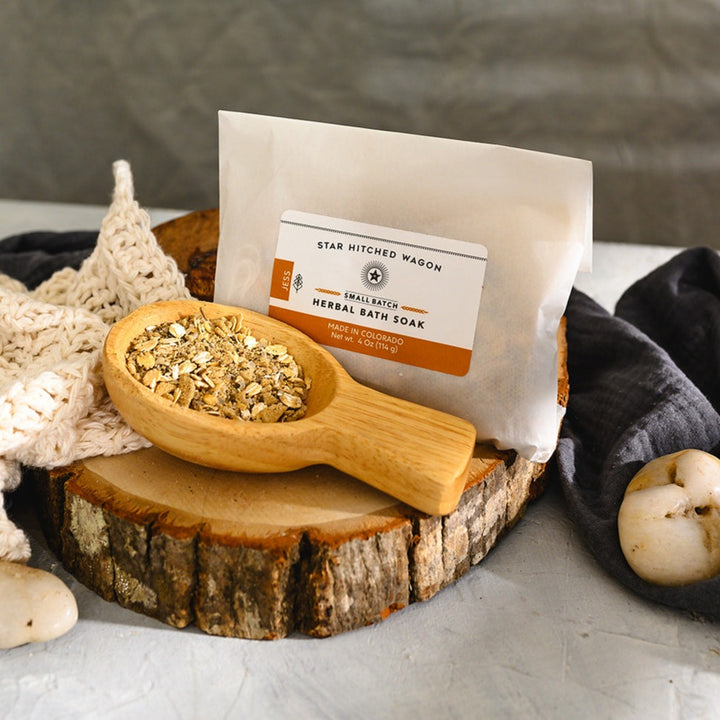 Wooden scoop filled with Jess Herbal Bath Soak next to full pouch on wooden slab next to a cream colored knit washcloth, a grey towel and pebbles.
