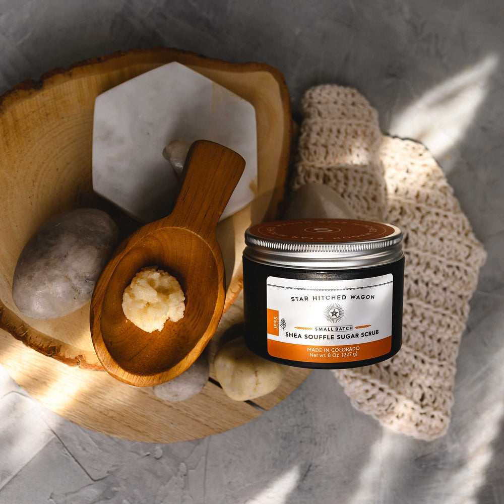 A jar of Jess 8 Ounce Shea Soufflé Sugar Scrub is displayed on a wooden surface, alongside a wooden spoon, a knit washcloth and small river stones.