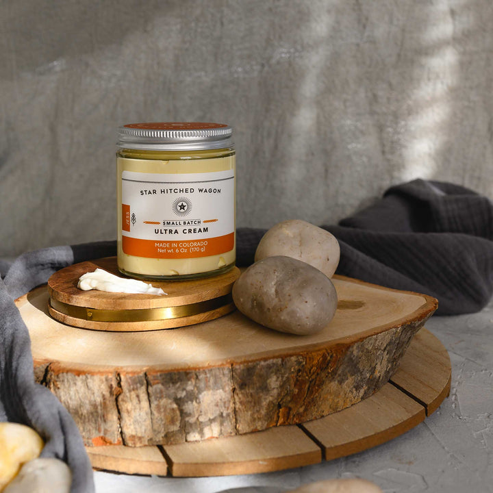 Jess Ultra Cream in a glass jar placed on a wooden platform, pebbles and grey towel in foreground and background.
