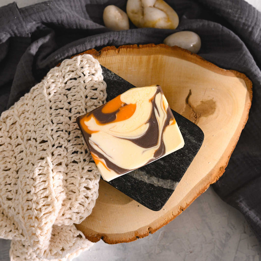 Jess essential oil soap bar with a swirl pattern of orange, brown, and cream colors, placed on a wooden surface with a cream colored knitted washcloth beside it.