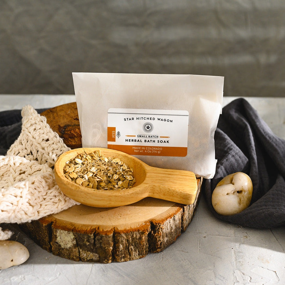 Wooden scoop filled with Jess Herbal Bath Soak next to full pouch on wooden slab next to a cream colored knit washcloth, a grey towel and pebbles.