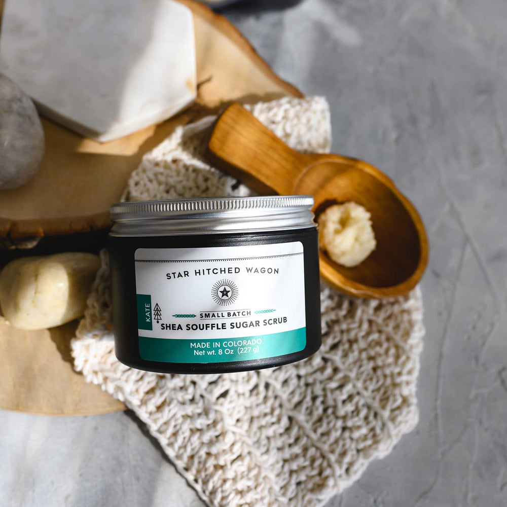 An 8 ounce container of Kate shea souffle sugar scrub is displayed with a wooden spoon showing the product, surrounded by natural elements like rocks and a knit washcloth, on a wooden surface.