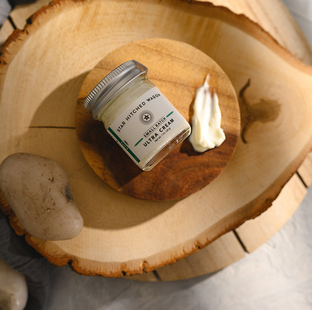 A small glass jar of Star Hitched Wagon Kate Ultra Cream lays on a round wooden slice, with a dollop of ultra cream next to jar.