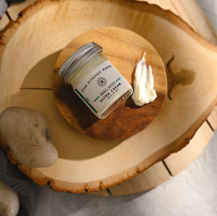 A small glass jar of Star Hitched Wagon Kate Ultra Cream lays on a round wooden slice, with a dollop of ultra cream next to jar.